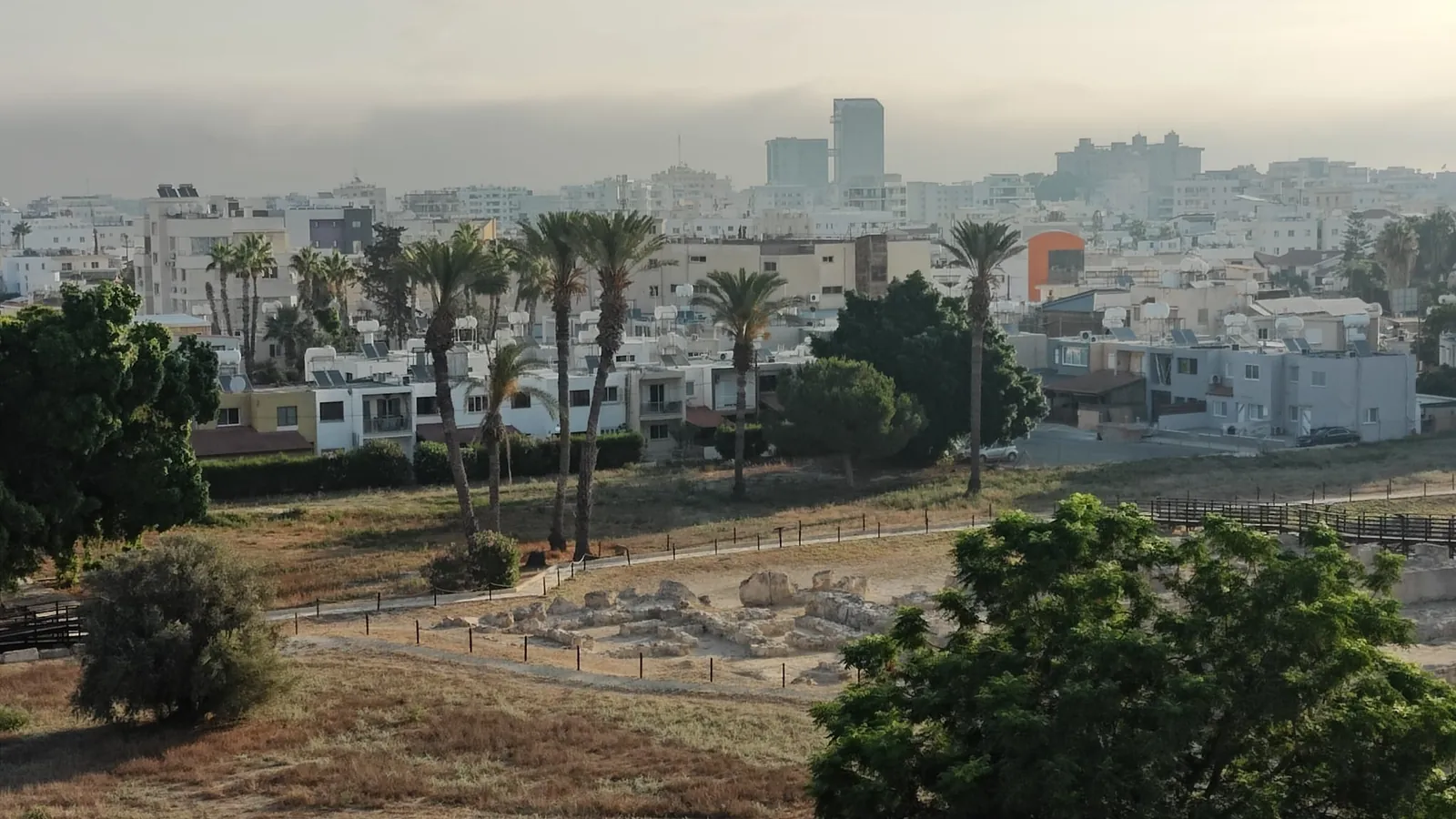 The excavated ruins of ancient Kition in the foreground, fenced and gridded; behind them the inhabited city of Larnaca — flats, palms, rooftops — continuing above the site.